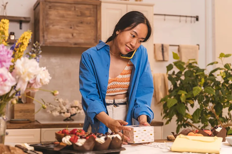 Pemilik usaha kecil menelepon supplier untuk negosiasi harga sambil mengemas pesanan di meja dapur