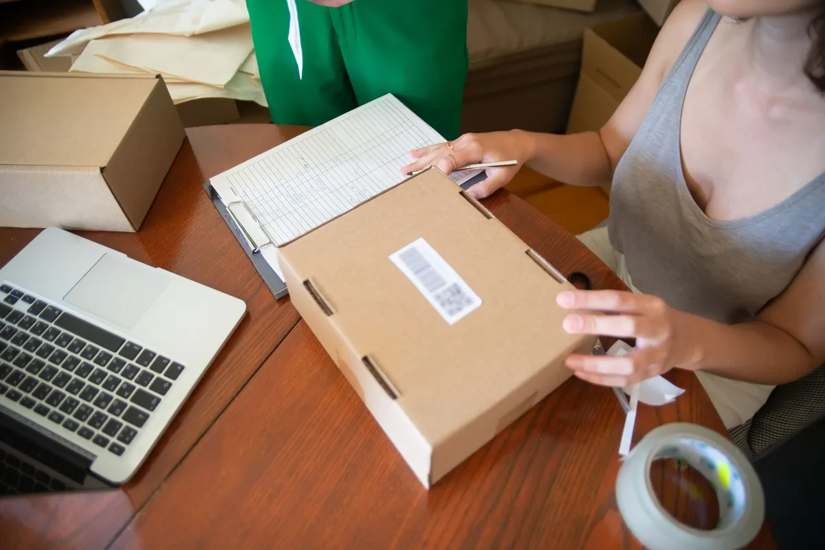 Penjual online menempelkan label barcode pada kotak paket di meja kayu, dengan laptop dan lakban di dekatnya