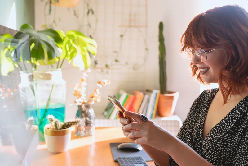 Wanita tersenyum melihat notifikasi di smartphone di ruang kerja rumahnya