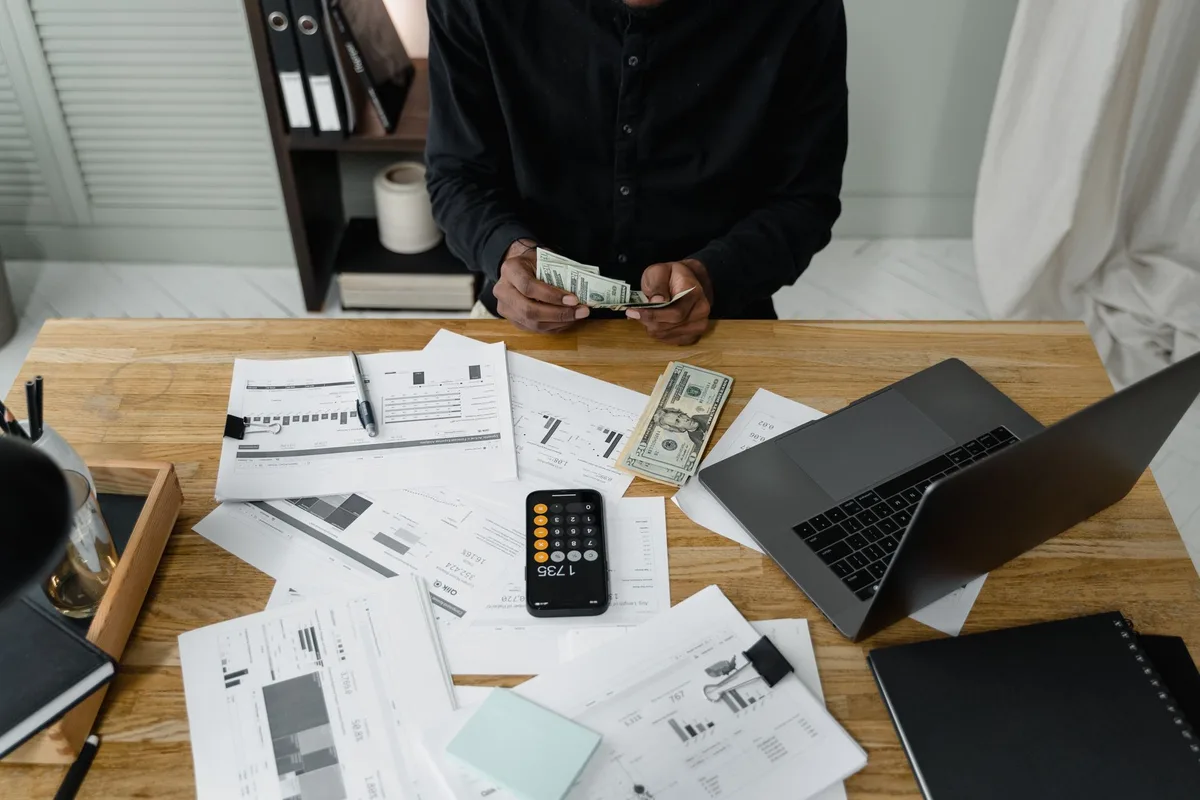 Person counting cash and reviewing a laptop budget spreadsheet at a desk